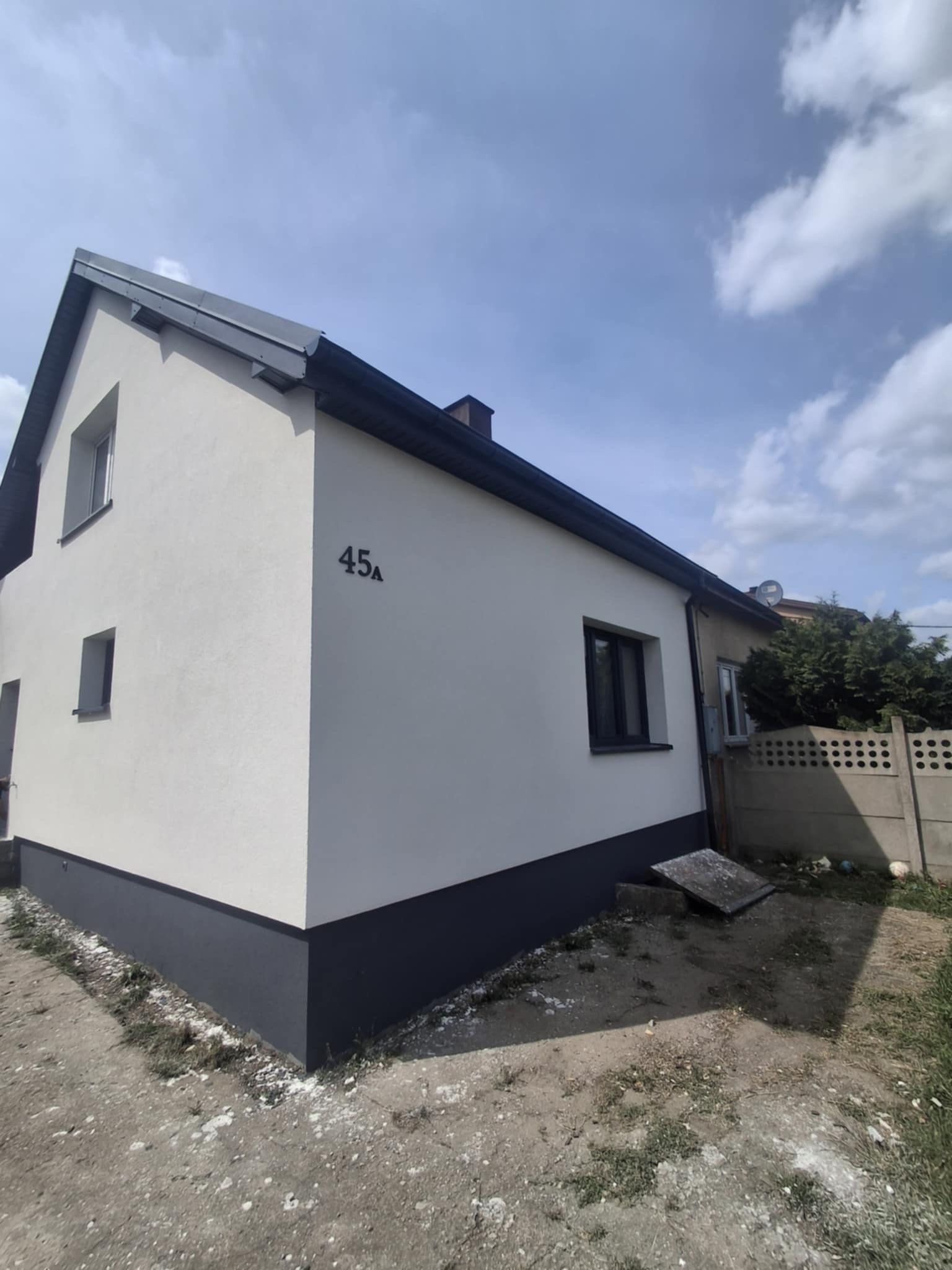 Odnowiona elewacja domu jednorodzinnego z numerem 45A, świeżo pomalowana na biało z szarym cokołem i obramowaniami okien, na tle błękitnego nieba z chmurami.