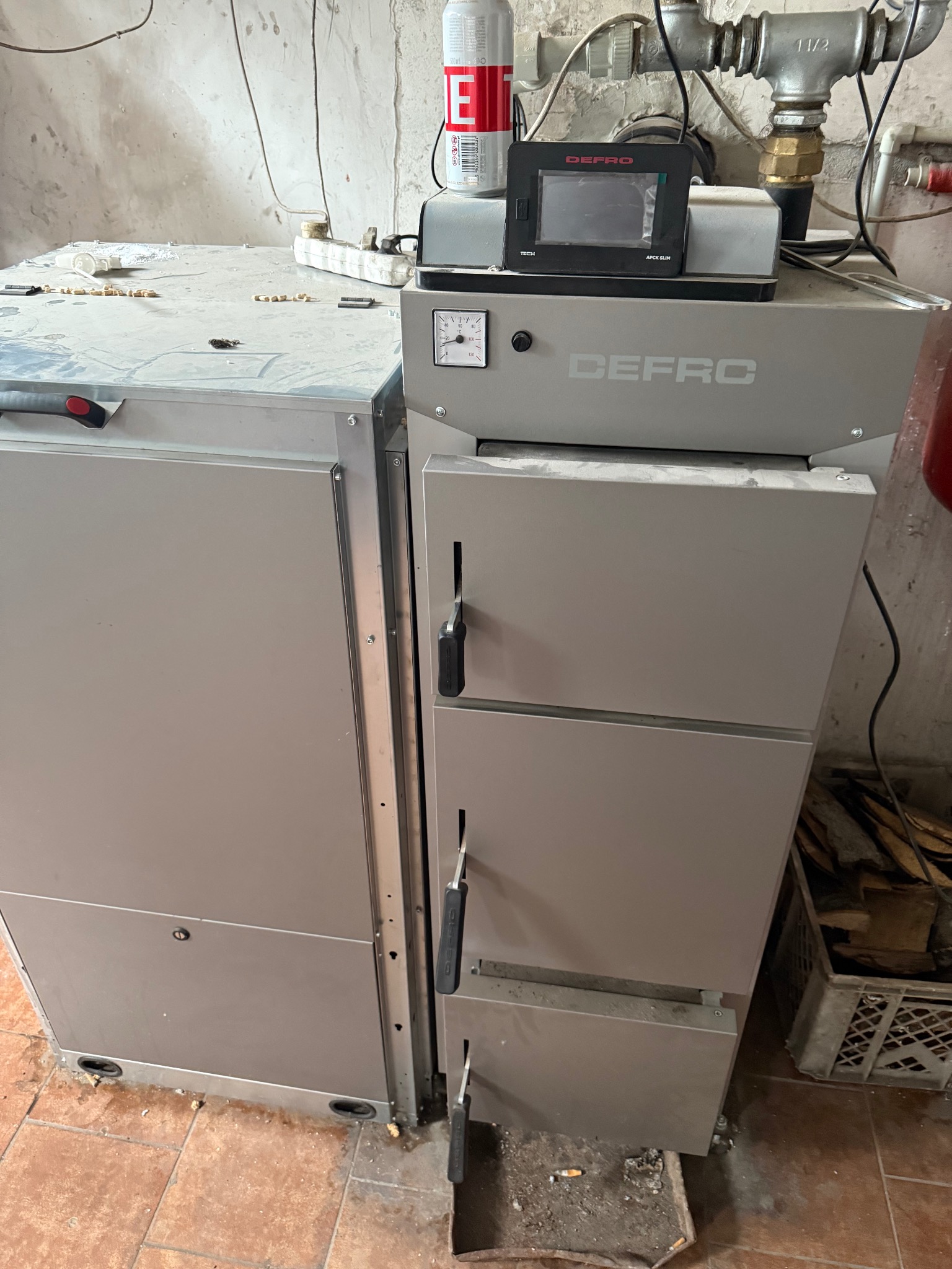Szary piec Defro z panelem sterowania i rurami, obok skrzynia z drewnem, na podłodze płytki. Widoczne ślady użytkowania i zabrudzenia.