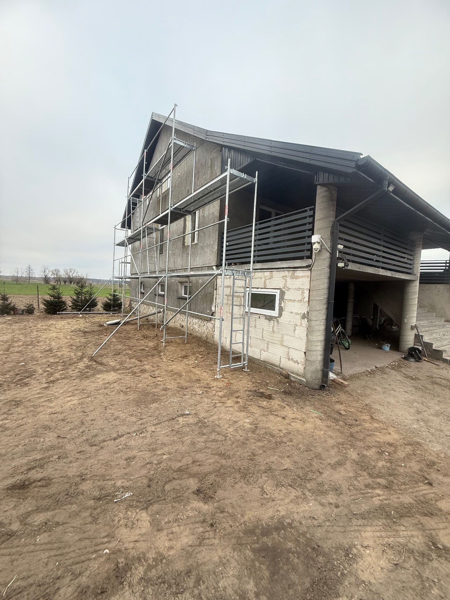 Dom w trakcie prac wykończeniowych z rusztowaniem. Widoczne surowe ściany z bloczków, częściowo wykończony balkon i teren wokół budynku.