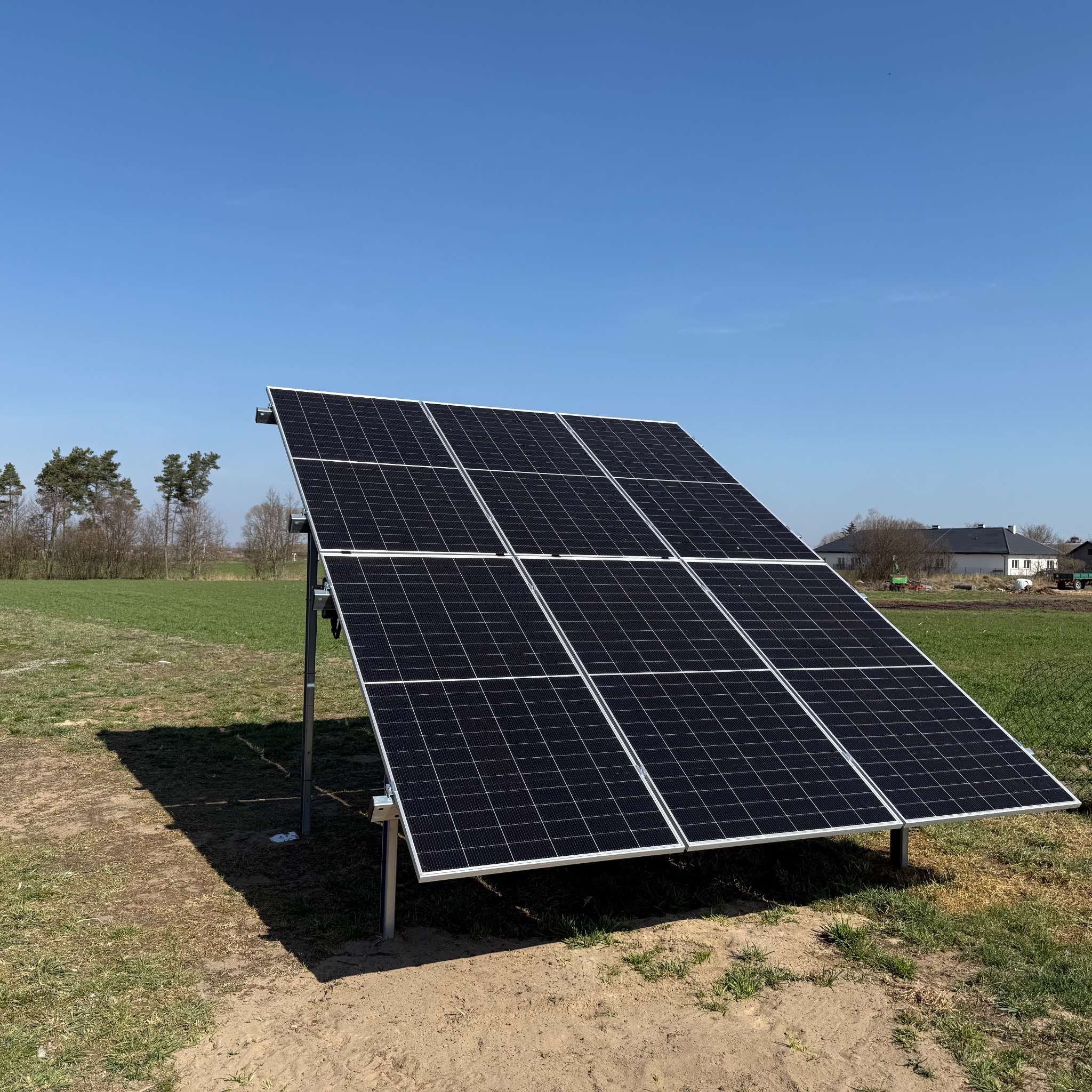 Panel słoneczny na łące w słoneczny dzień. Widoczne moduły fotowoltaiczne, konstrukcja wsporcza i trawa. W tle budynek i drzewa. Czyste niebo.