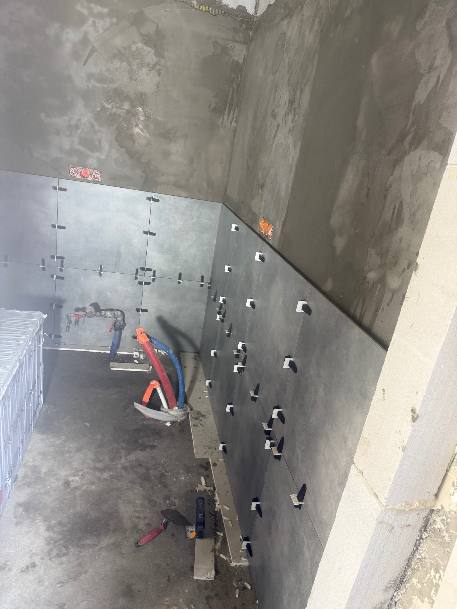 Wnętrze w trakcie prac wykończeniowych: szare płytki na ścianach z krzyżykami dystansowymi, widoczne narzędzia glazurnicze i instalacje. Surowy stan ścian i podłogi.