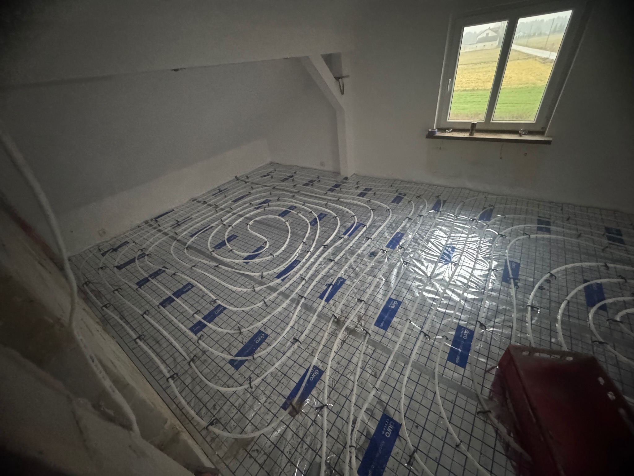 Białe rury ogrzewania podłogowego ułożone na siatce w surowym pokoju na poddaszu z oknem. Widok na zewnątrz przez okno.