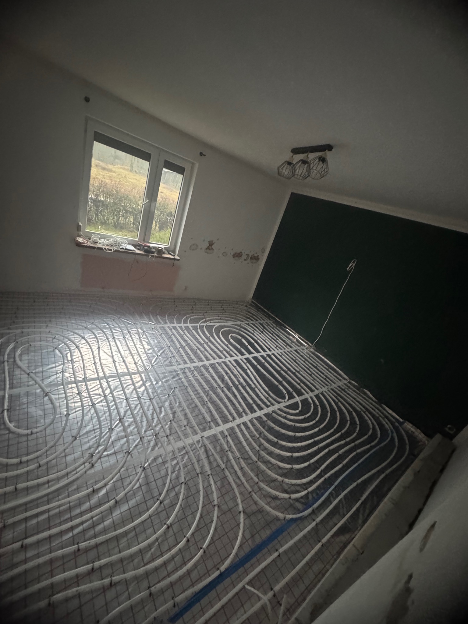 Białe rury ogrzewania podłogowego ułożone na siatce w pokoju z oknem i ciemną ścianą. Widoczny etap prac instalacyjnych przed wylaniem posadzki.