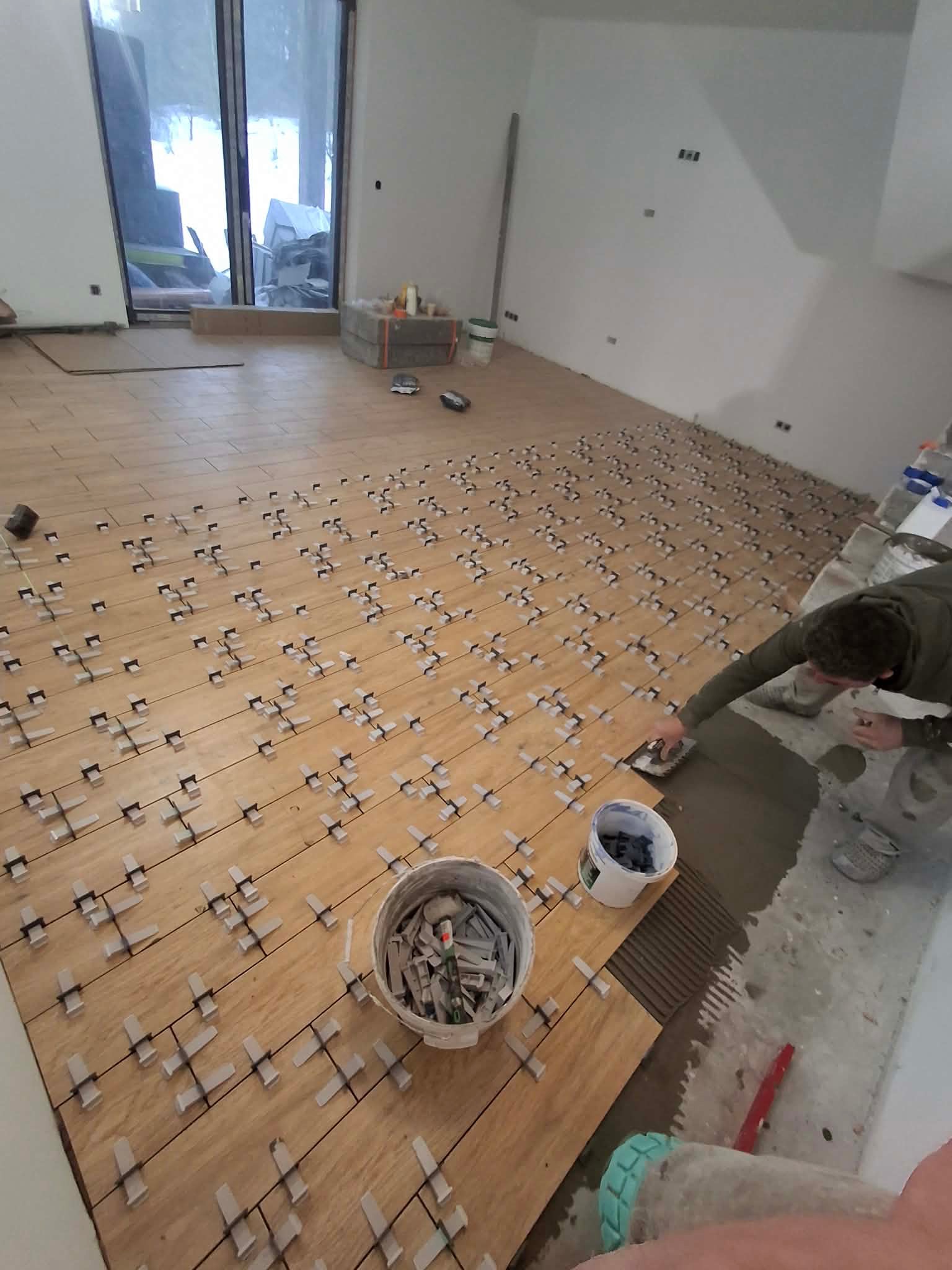 Układanie płytek podłogowych w jasnym odcieniu drewna, z użyciem krzyżyków dystansowych. Widoczny fragment pomieszczenia z dużym oknem i pracownik w trakcie pracy.