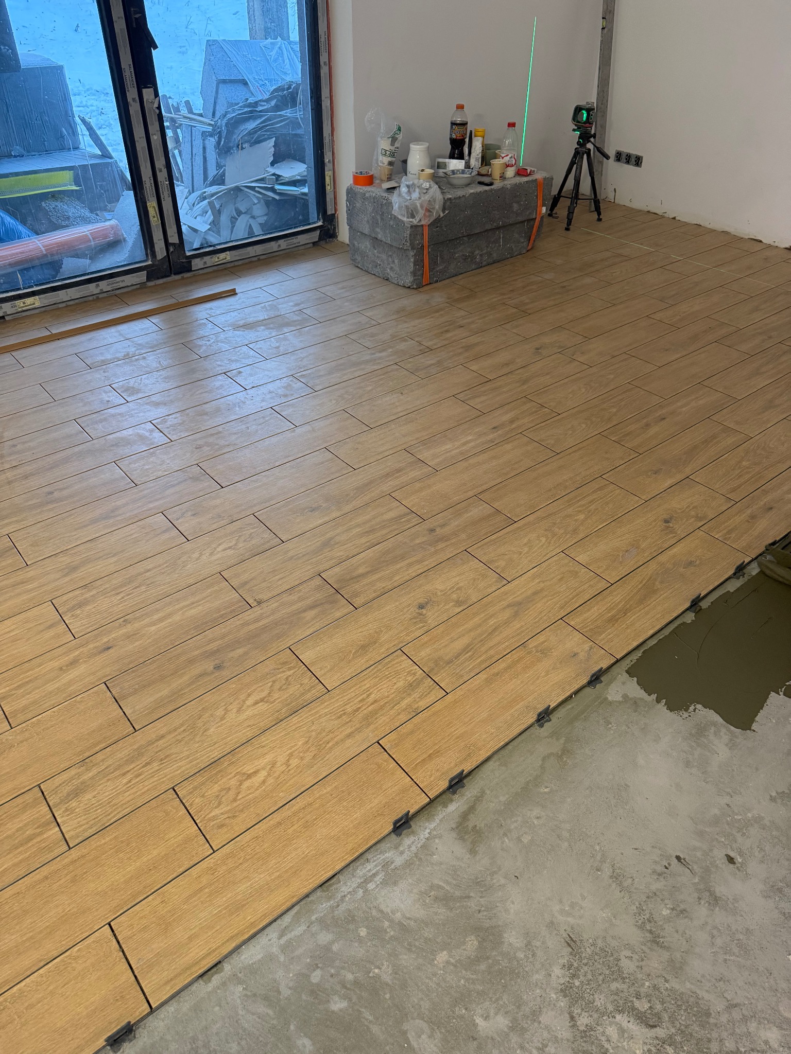Układanie podłogi z płytek imitujących drewno, widoczny laser krzyżowy, klej i system poziomowania. Częściowo wykończona podłoga w jasnym wnętrzu.