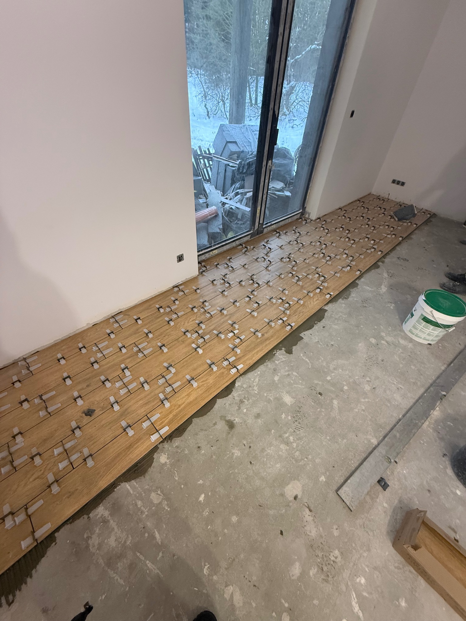 Układanie drewnopodobnych płytek podłogowych z użyciem krzyżyków dystansowych przy dużym oknie. Widoczne narzędzia i wiadro z klejem. Etap prac wykończeniowych.