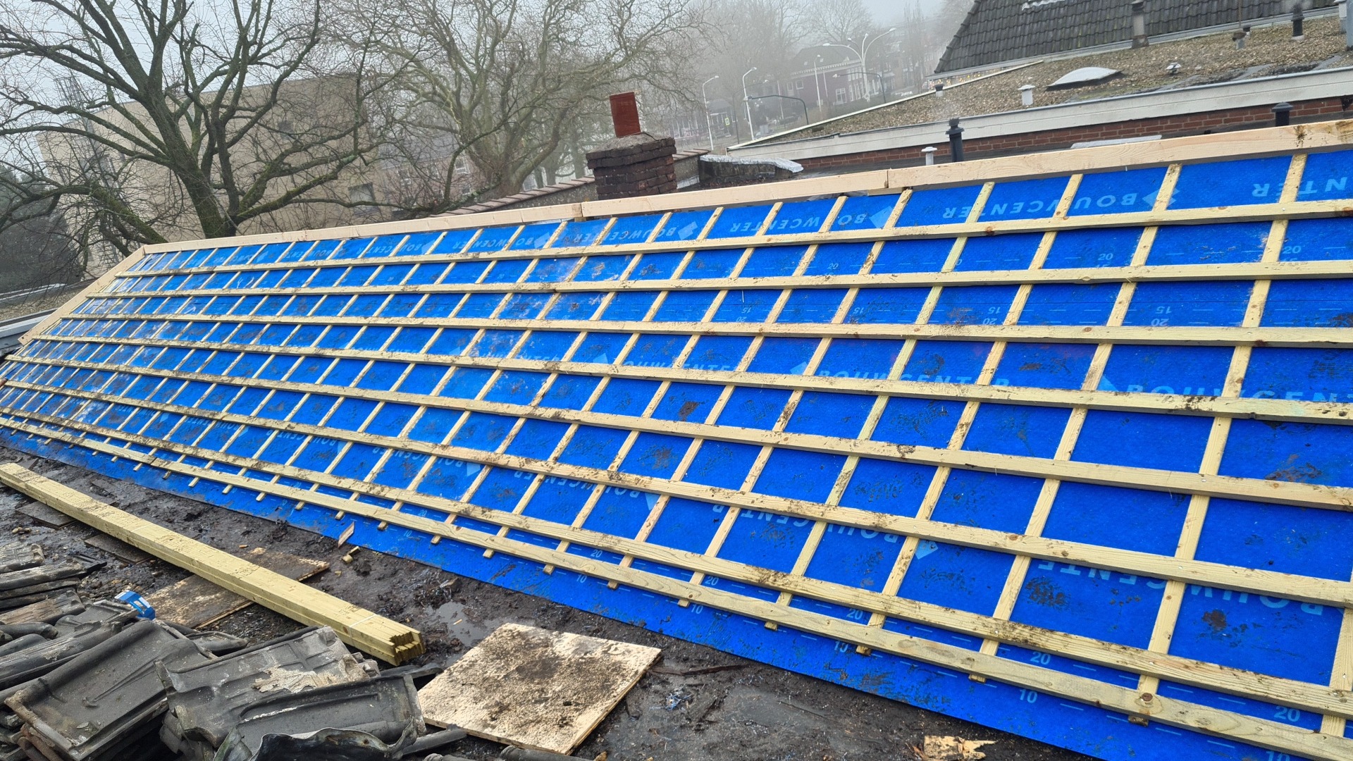Dach w trakcie przygotowania do położenia pokrycia: widoczne drewniane łaty na niebieskiej membranie dachowej z widocznymi napisami, w tle komin i zabudowania.
