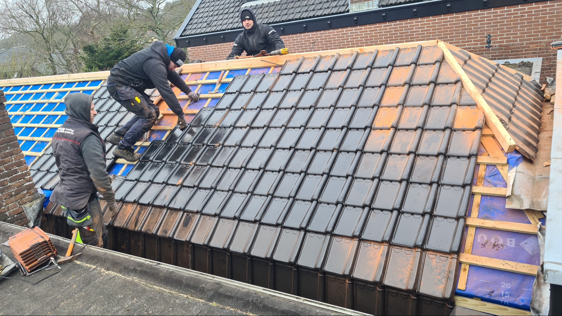 Trzech dekarzy pracuje na dachu, układając czarne dachówki na łatach, widoczne zabezpieczenie folią, część dachu jest już pokryta, a część przygotowana do dalszego układania.