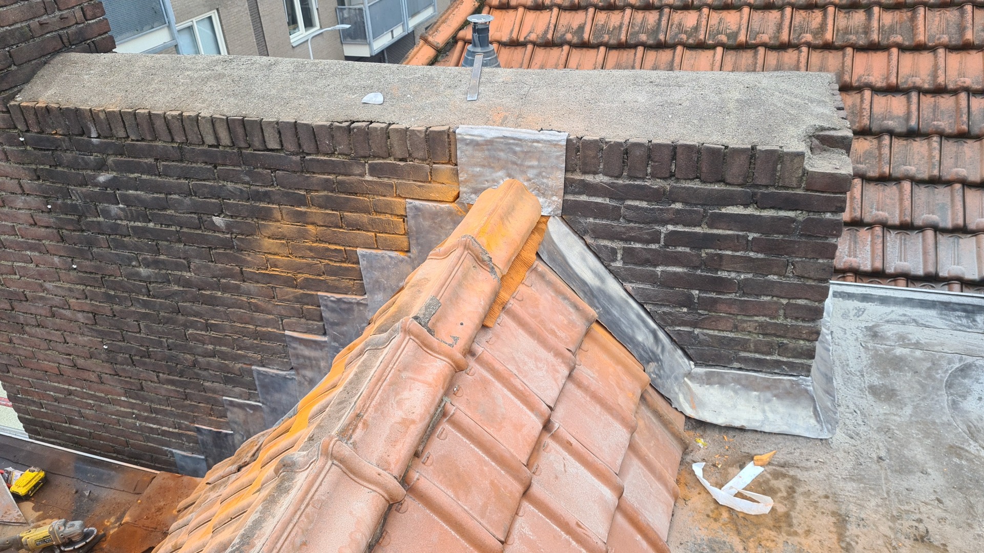 Widok z góry na fragment dachu z pomarańczową dachówką ceramiczną i komin z cegły z obróbkami blacharskimi z blachy, widoczne narzędzia pracy.