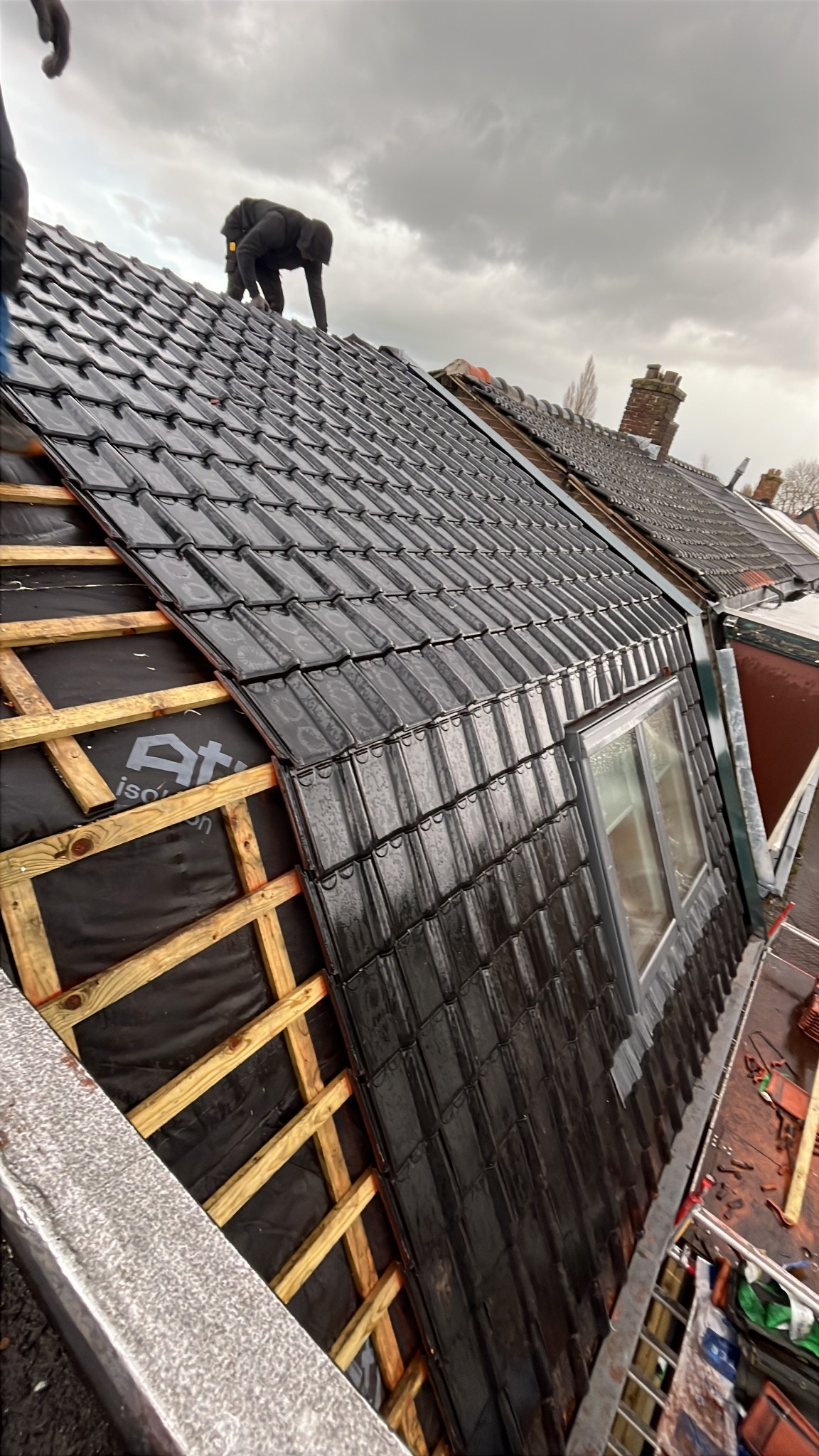 Montaż czarnej dachówki karpiówki na łatach z desek, widok z góry na fragment dachu z oknem połaciowym, dekarz na dachu, pochmurne niebo.