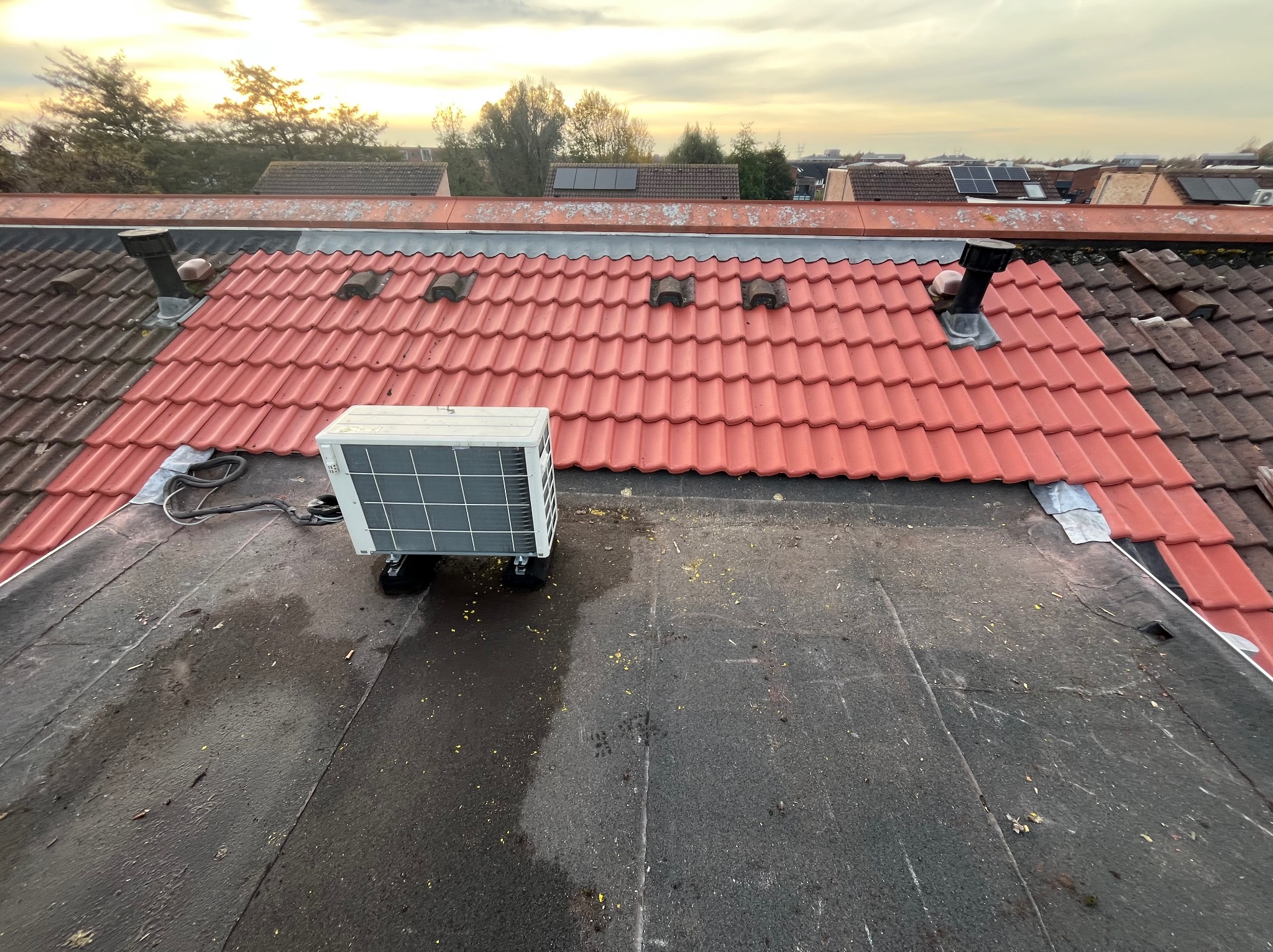 Częściowo odnowiony dach z czerwoną dachówką, widoczny fragment starego dachu i klimatyzator na płaskiej części dachu. Widok z góry, w tle zabudowa i drzewa.
