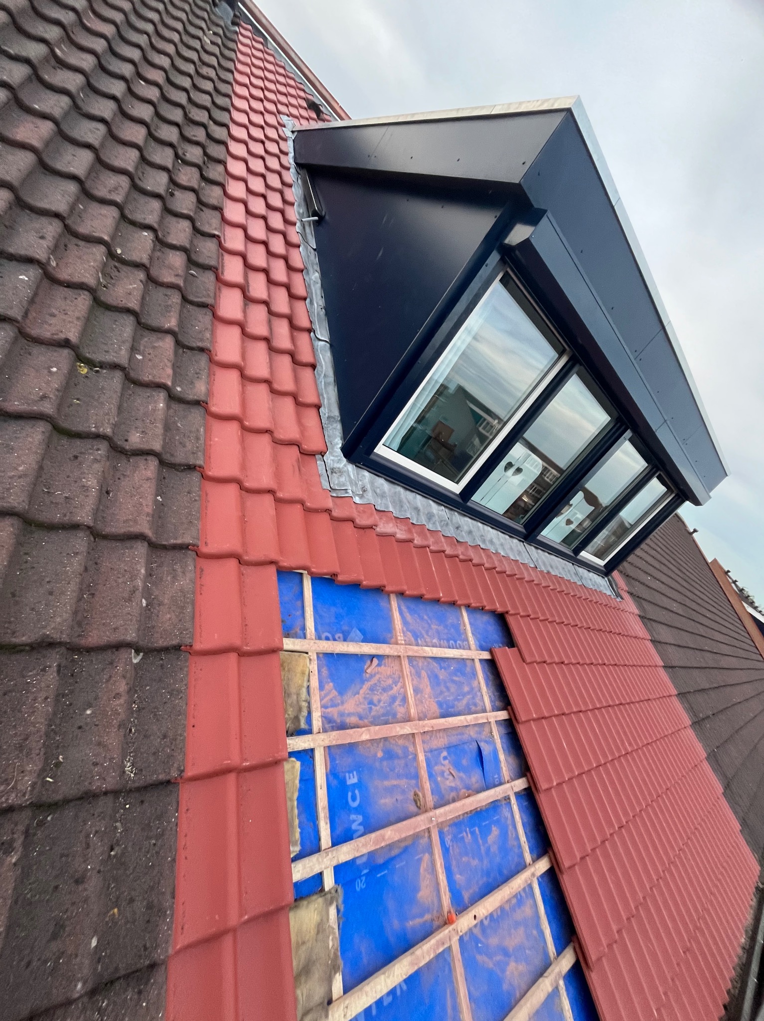 Częściowo wymienione czerwone dachówki kontrastują z ciemnymi, starymi. Nowe okno dachowe z czarną obudową. Widoczna izolacja pod dachówkami.