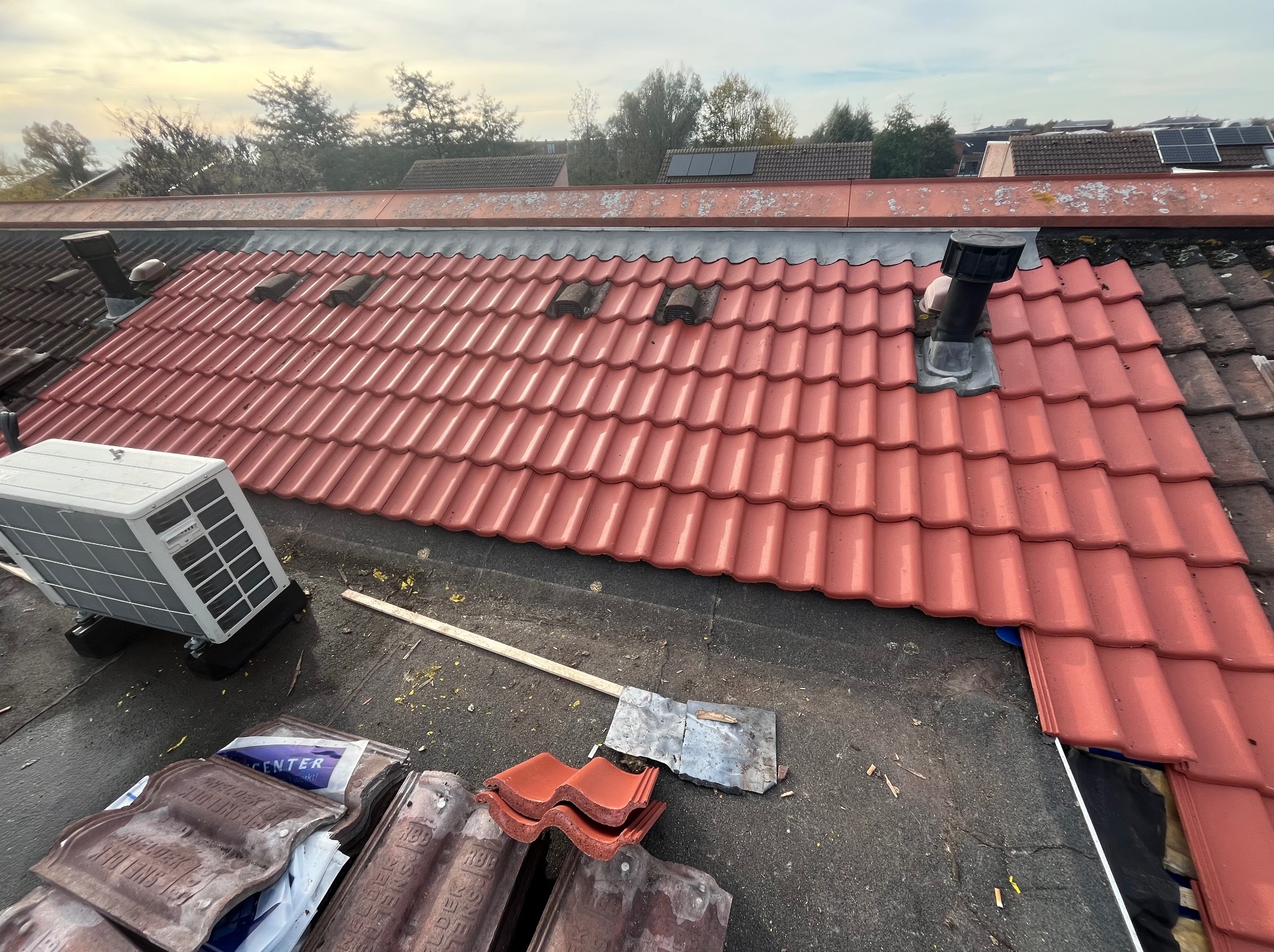 Częściowo pokryty dach czerwoną dachówką, widoczne narzędzia, wentylacja i klimatyzator. Fragment dachu pokryty starą dachówką. Prace dekarskie w toku.