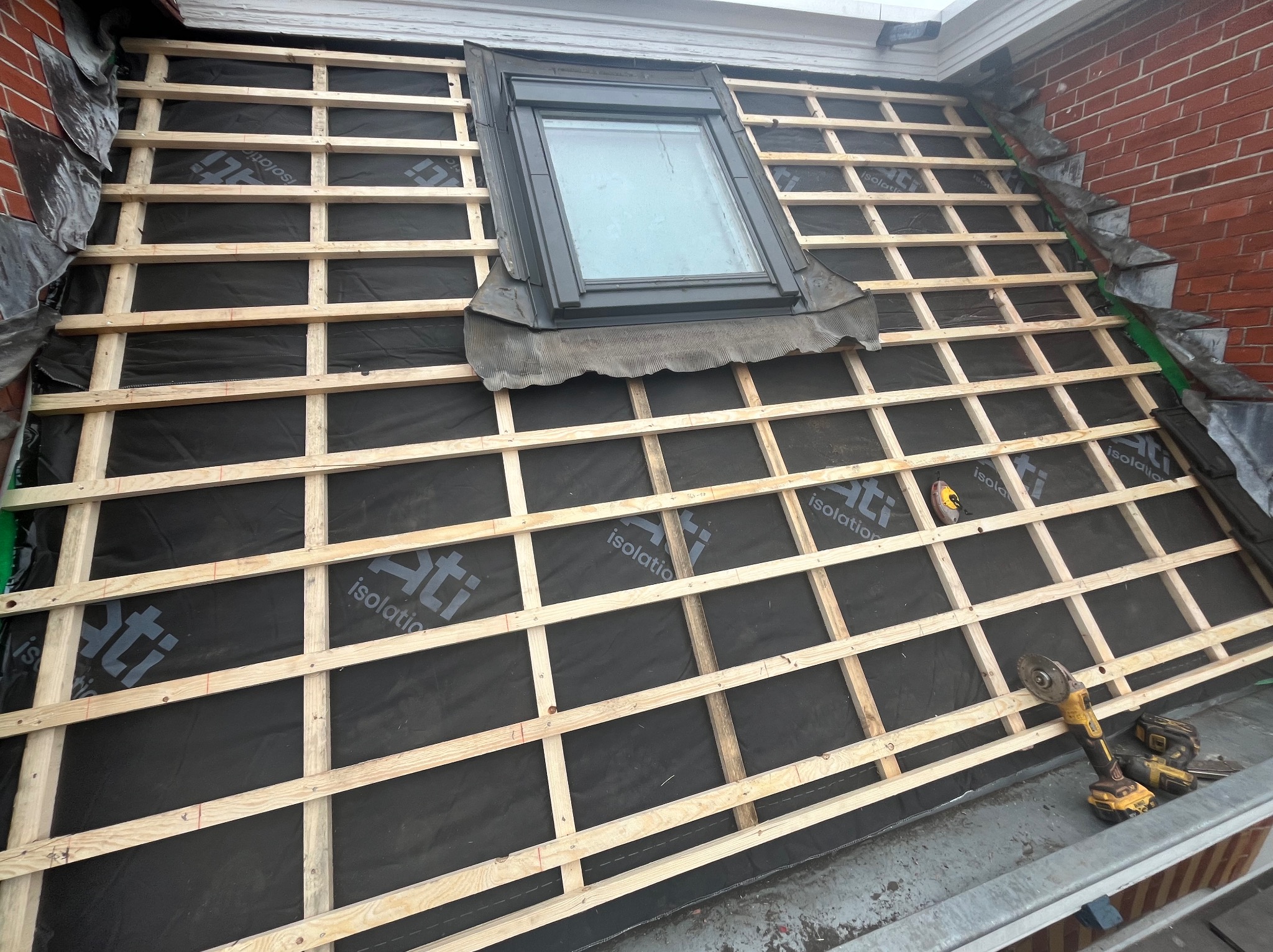 Montaż łat na membranie dachowej z oknem połaciowym. Widoczne narzędzia: szlifierka kątowa i miara zwijana. Etap przygotowania pod krycie dachu.