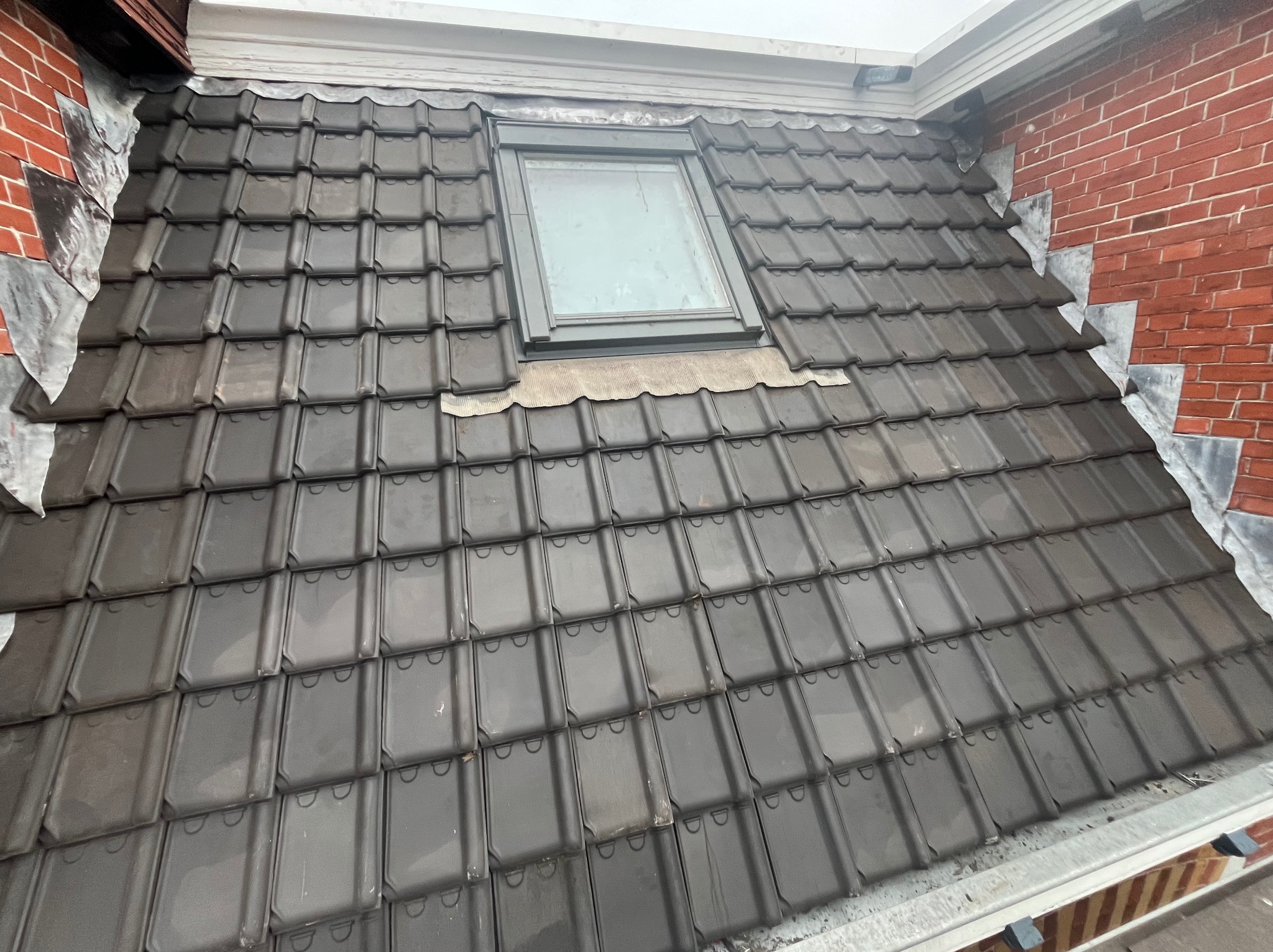 Ukończony dach z ciemnej dachówki ceramicznej, z oknem dachowym i obróbkami blacharskimi przy kominie z czerwonej cegły. Widok z góry, lekki kąt.