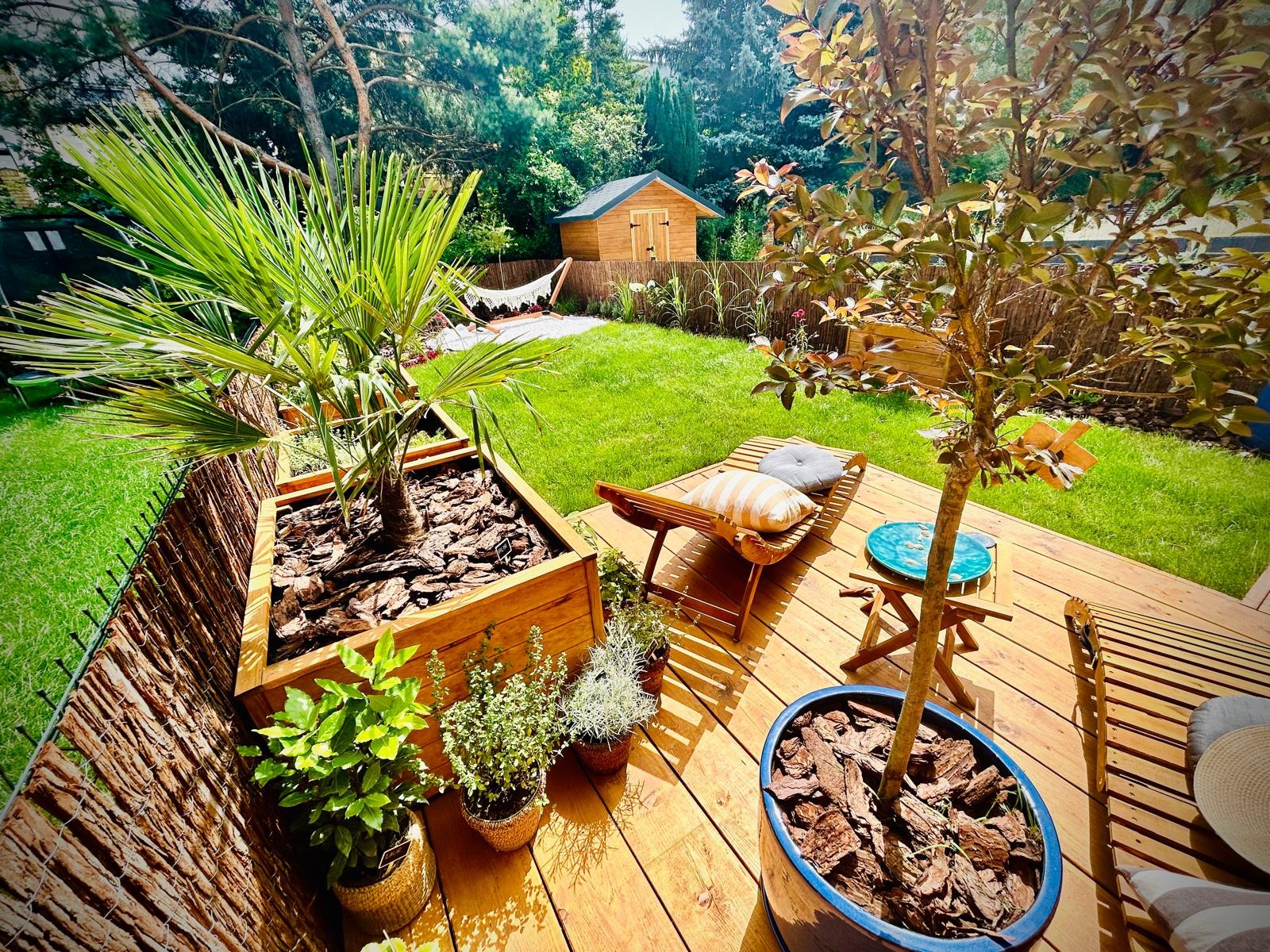 Urokliwy ogród z drewnianym tarasem, palmą w dużej donicy, leżakiem z poduszkami, stolikiem i hamakiem w tle, otoczony zielenią i drewnianą chatką.