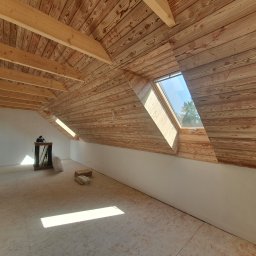 PROFIDOM Firma Ogólnobudowlana - Poddasze z drewnianą boazerią na skosach dachu, okno dachowe wpuszczające światło, surowa podłoga, przygotowane do dalszych prac wykończeniowych.