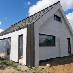 PROFIDOM Firma Ogólnobudowlana - Nowoczesny dom jednorodzinny w trakcie prac wykończeniowych elewacji, widoczne fragmenty ocieplenia, świeżo nałożona warstwa tynku, okna zabezpieczone folią, na ziemi wiadro z farbą, pracownik w tle.