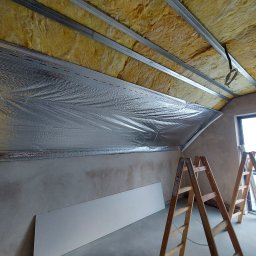 PROFIDOM Firma Ogólnobudowlana - Poddasze w trakcie izolacji: widoczna wełna mineralna pomiędzy metalowymi profilami, folia aluminiowa oraz dwie drewniane drabiny opierające się o ścianę, w tle okno.