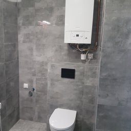 PROFIDOM Firma Ogólnobudowlana - Nowoczesna łazienka z szarymi płytkami imitującymi kamień, wiszącą toaletą i białym piecem gazowym z miedzianymi rurami.