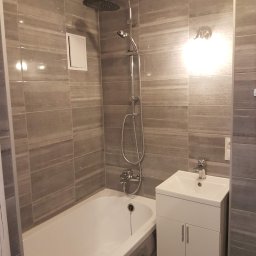 PROFIDOM Firma Ogólnobudowlana - Nowoczesna łazienka z wanną, umywalką z szafką i szarą, poziomą glazurą imitującą kamień, oświetlona punktowym światłem.