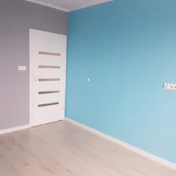 Pomieszczenie po malowaniu ścian na szaro i błękitno, z białą, nowoczesną stolarką drzwiową i jasnymi panelami podłogowymi.