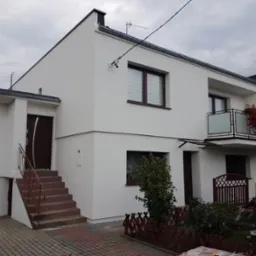 Dwupiętrowy dom jednorodzinny z ocieploną i pomalowaną na biało elewacją, balkon z kwiatami, brązowe okna i drzwi, schody wejściowe z metalową balustradą, kostka brukowa na podjeździe, pochmurne...