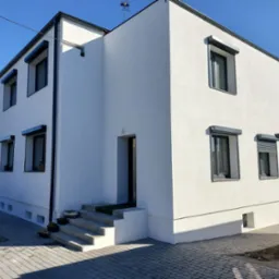 Nowoczesny, dwupiętrowy dom z białą elewacją i szarymi roletami zewnętrznymi, widoczny z boku, z kostką brukową na podjeździe i anteną na dachu.