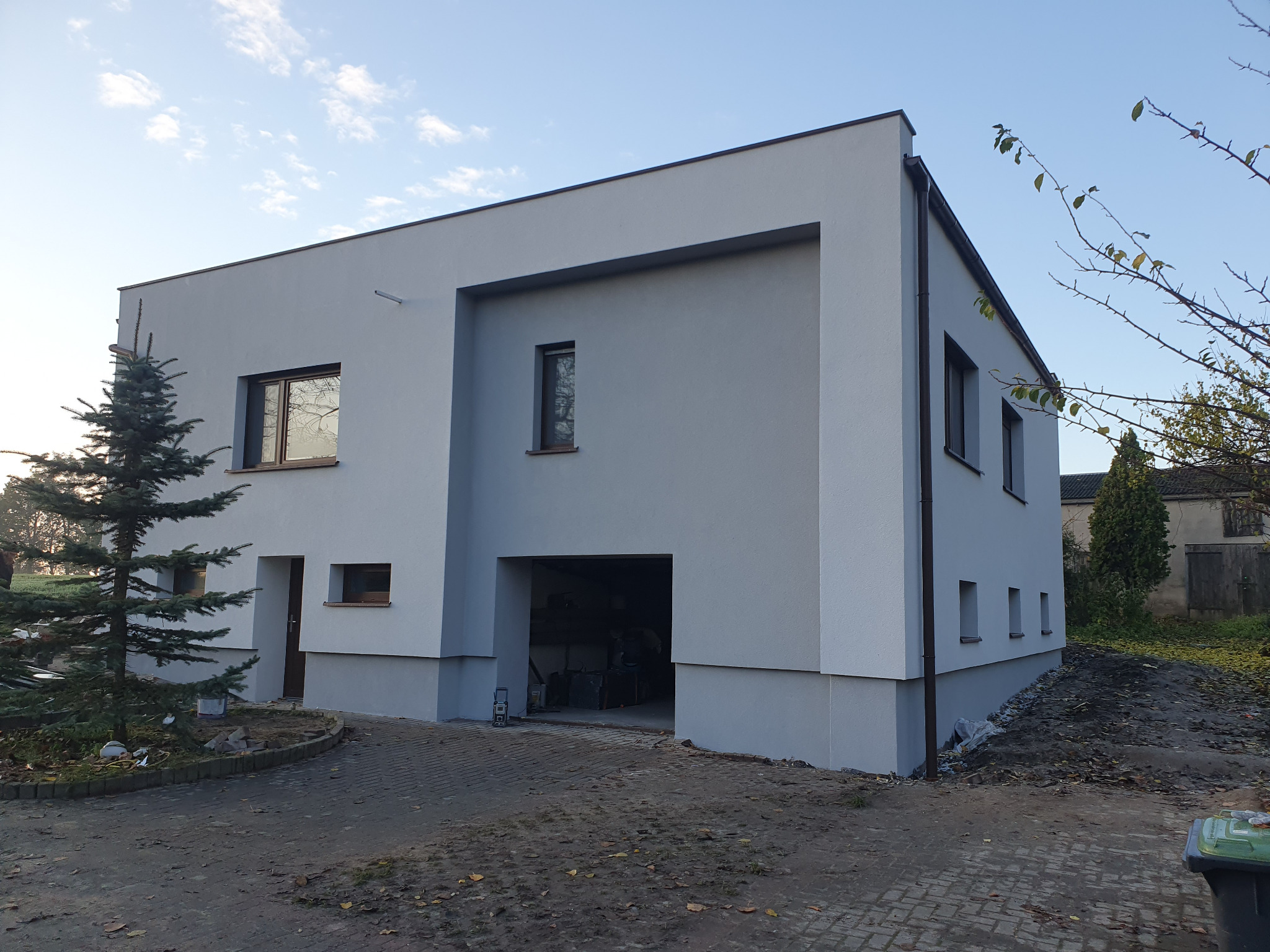 Nowoczesny dom jednorodzinny z szarą elewacją i brązowymi ramami okiennymi, widok z zewnątrz na garaż, teren wokół domu częściowo zagospodarowany, widoczna mała choinka i pozostałości po budowie.