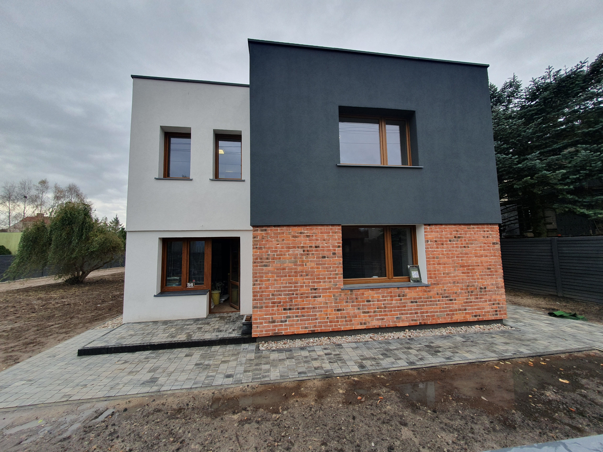 Nowoczesny dom z elewacją w dwóch kolorach: biały i grafitowy, z elementami z cegły klinkierowej, kostka brukowa przed wejściem, pochmurne niebo.