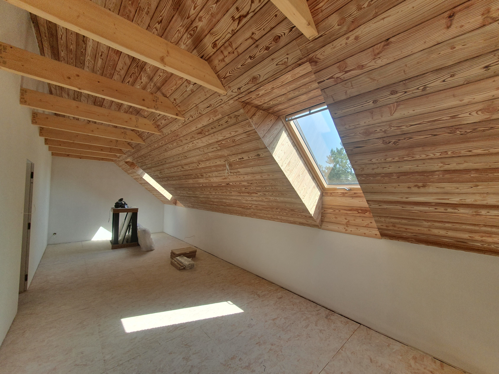 Poddasze z drewnianą boazerią na skosach dachu, okno dachowe wpuszczające światło, surowa podłoga, przygotowane do dalszych prac wykończeniowych.