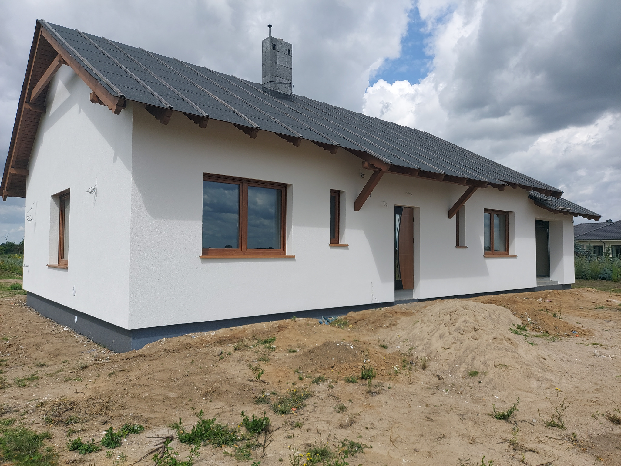 Świeżo otynkowany dom jednorodzinny z brązowymi oknami i ciemną podmurówką, widok z boku na tle pochmurnego nieba, teren wokół domu jeszcze niezagospodarowany.