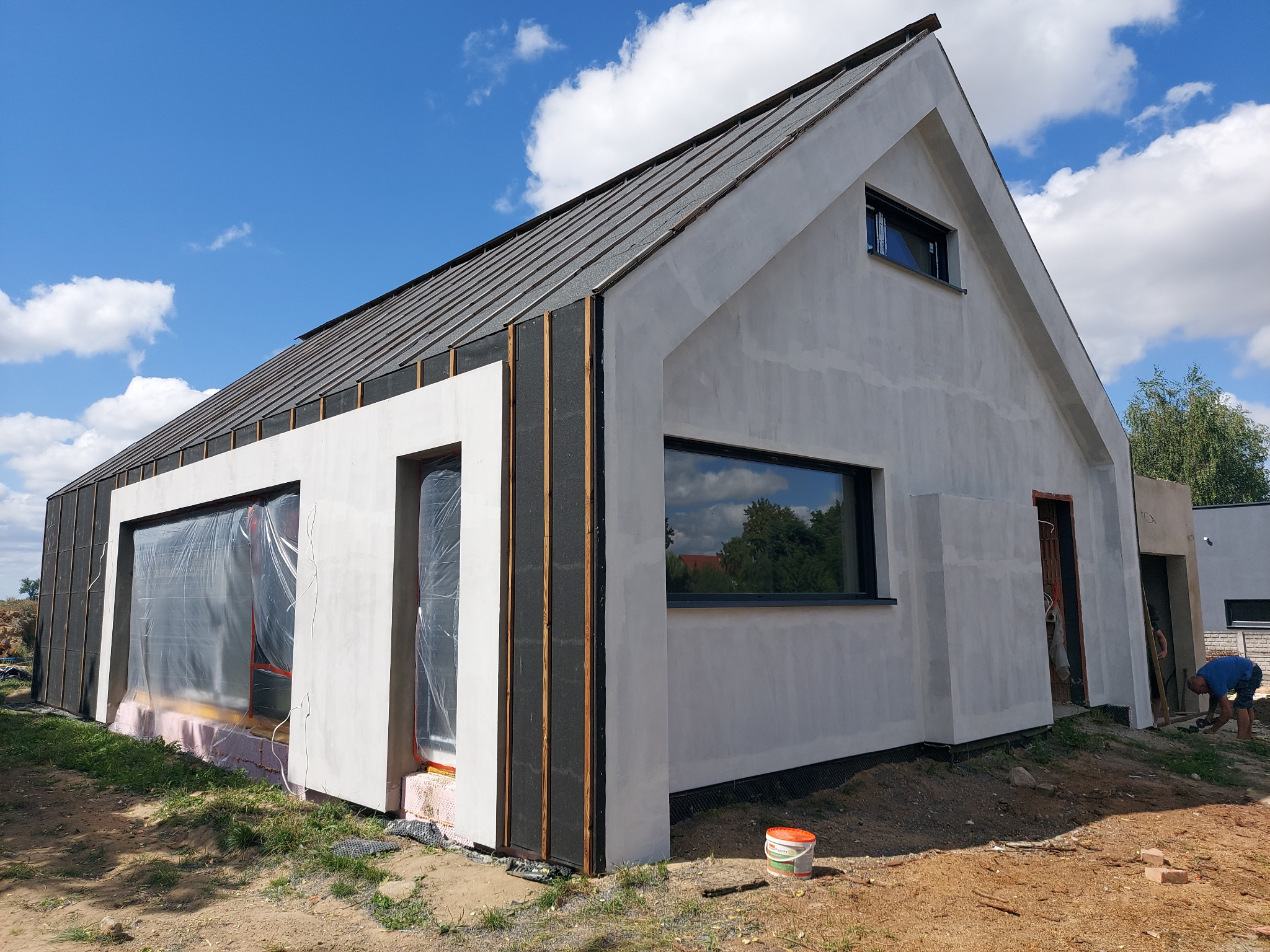 Nowoczesny dom jednorodzinny w trakcie prac wykończeniowych elewacji, widoczne fragmenty ocieplenia, świeżo nałożona warstwa tynku, okna zabezpieczone folią, na ziemi wiadro z farbą, pracownik w tle.