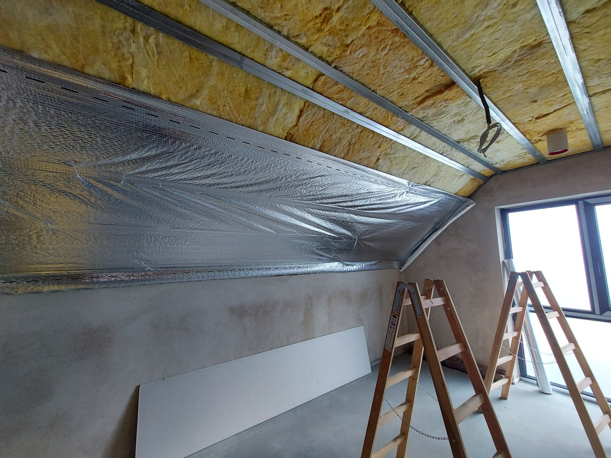 Poddasze w trakcie izolacji: widoczna wełna mineralna pomiędzy metalowymi profilami, folia aluminiowa oraz dwie drewniane drabiny opierające się o ścianę, w tle okno.