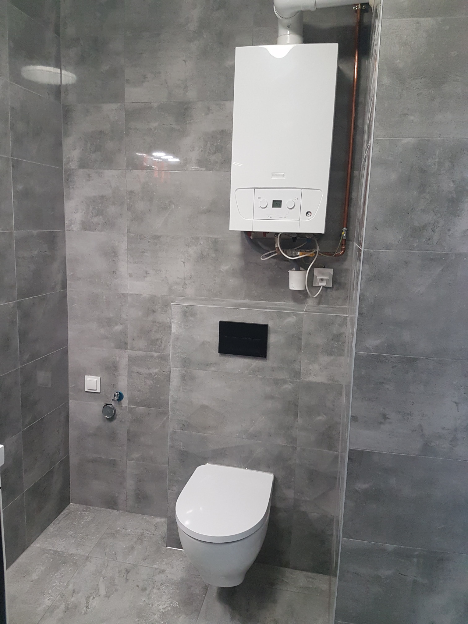 Nowoczesna łazienka z szarymi płytkami imitującymi kamień, wiszącą toaletą i białym piecem gazowym z miedzianymi rurami.