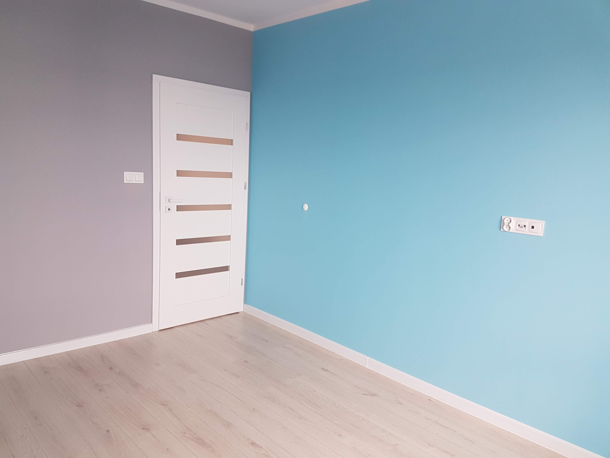 Pomieszczenie po malowaniu ścian na szaro i błękitno, z białą, nowoczesną stolarką drzwiową i jasnymi panelami podłogowymi.