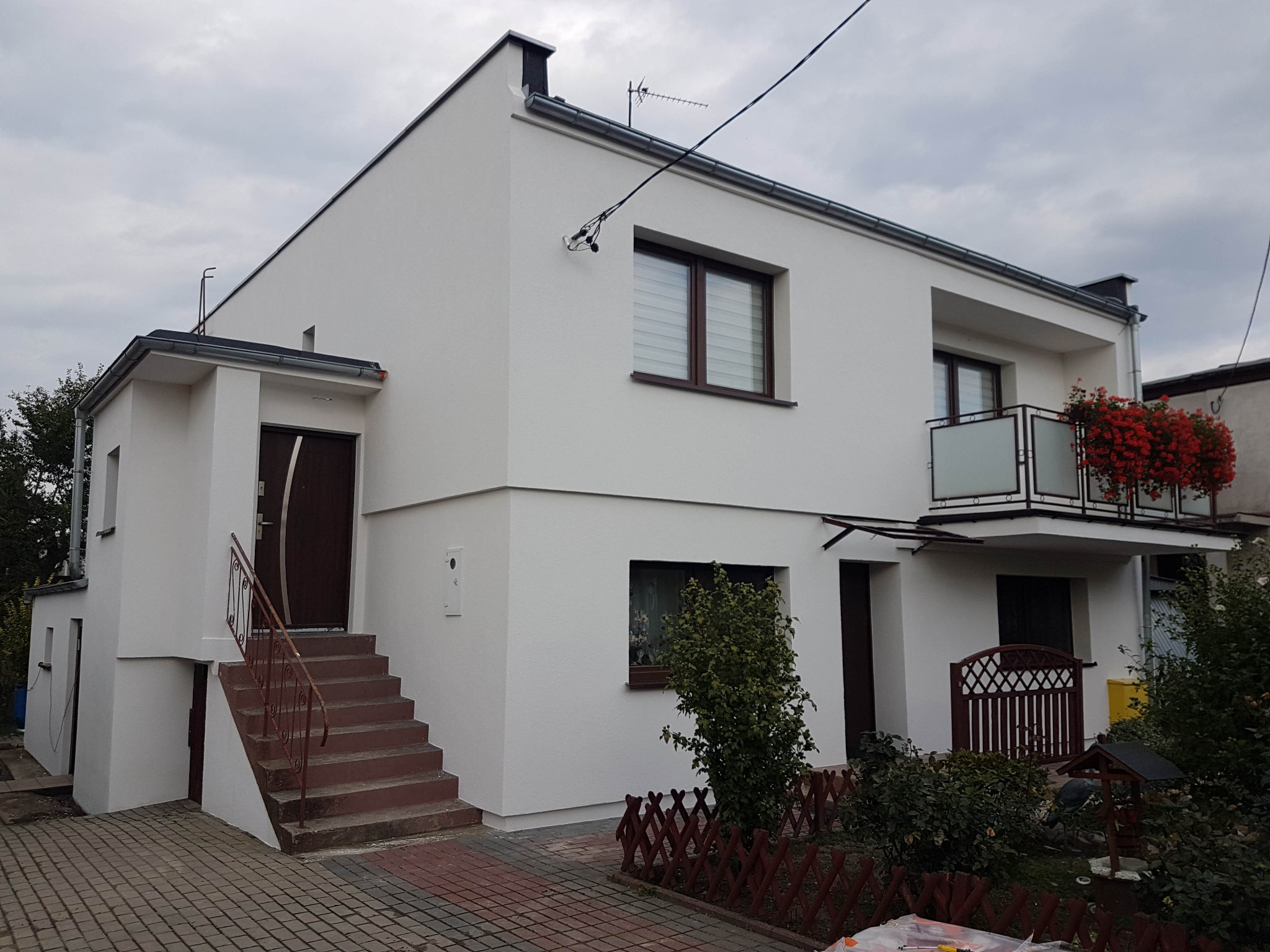 Dwupiętrowy dom jednorodzinny z ocieploną i pomalowaną na biało elewacją, balkon z kwiatami, brązowe okna i drzwi, schody wejściowe z metalową balustradą, kostka brukowa na podjeździe, pochmurne...