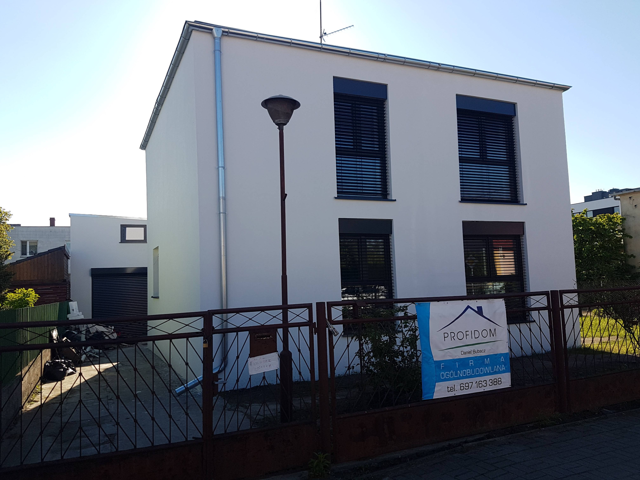 Nowoczesny, dwukondygnacyjny dom z białą elewacją i czarnymi roletami zewnętrznymi, widoczny zza metalowego ogrodzenia z reklamą firmy budowlanej. Na elewacji widoczne rury spustowe, a przed domem...