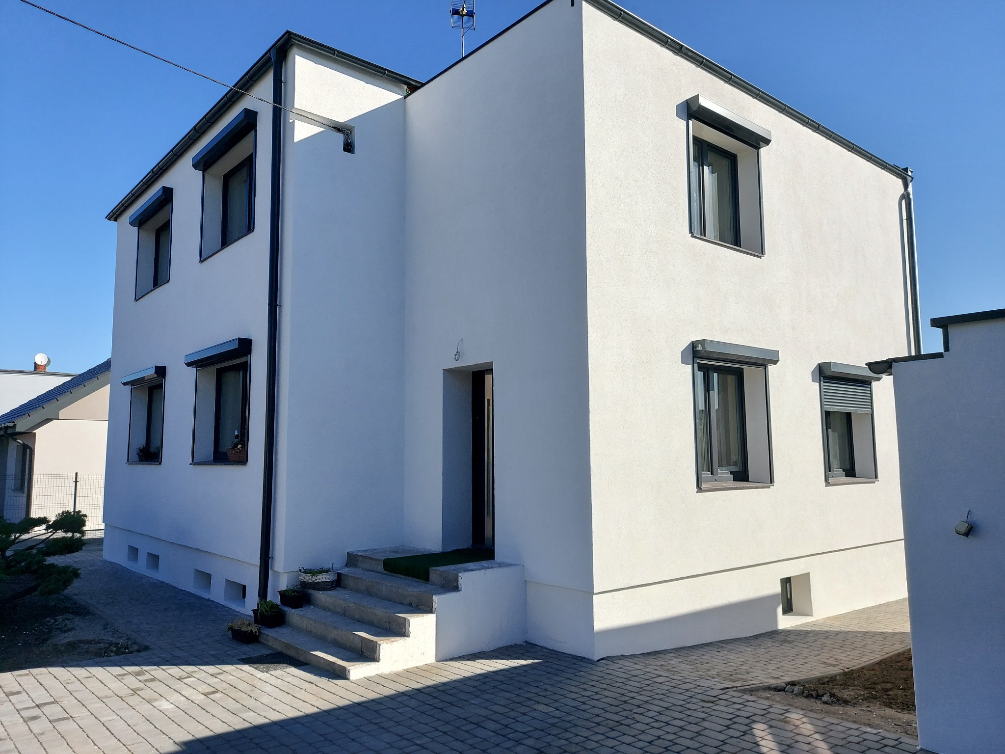 Nowoczesny, dwupiętrowy dom z białą elewacją i szarymi roletami zewnętrznymi, widoczny z boku, z kostką brukową na podjeździe i anteną na dachu.