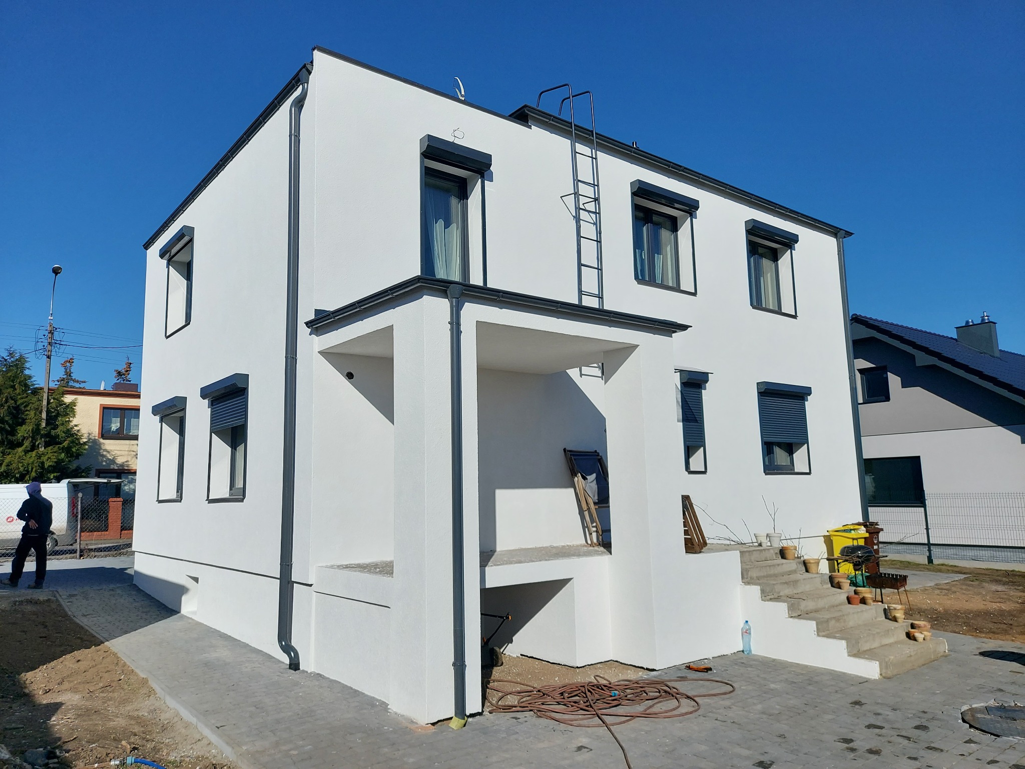 Nowoczesny, dwukondygnacyjny dom z białą elewacją, szarymi oknami z roletami zewnętrznymi, rynnami i schodami wejściowymi, na tle błękitnego nieba, z widocznymi elementami otoczenia w trakcie...