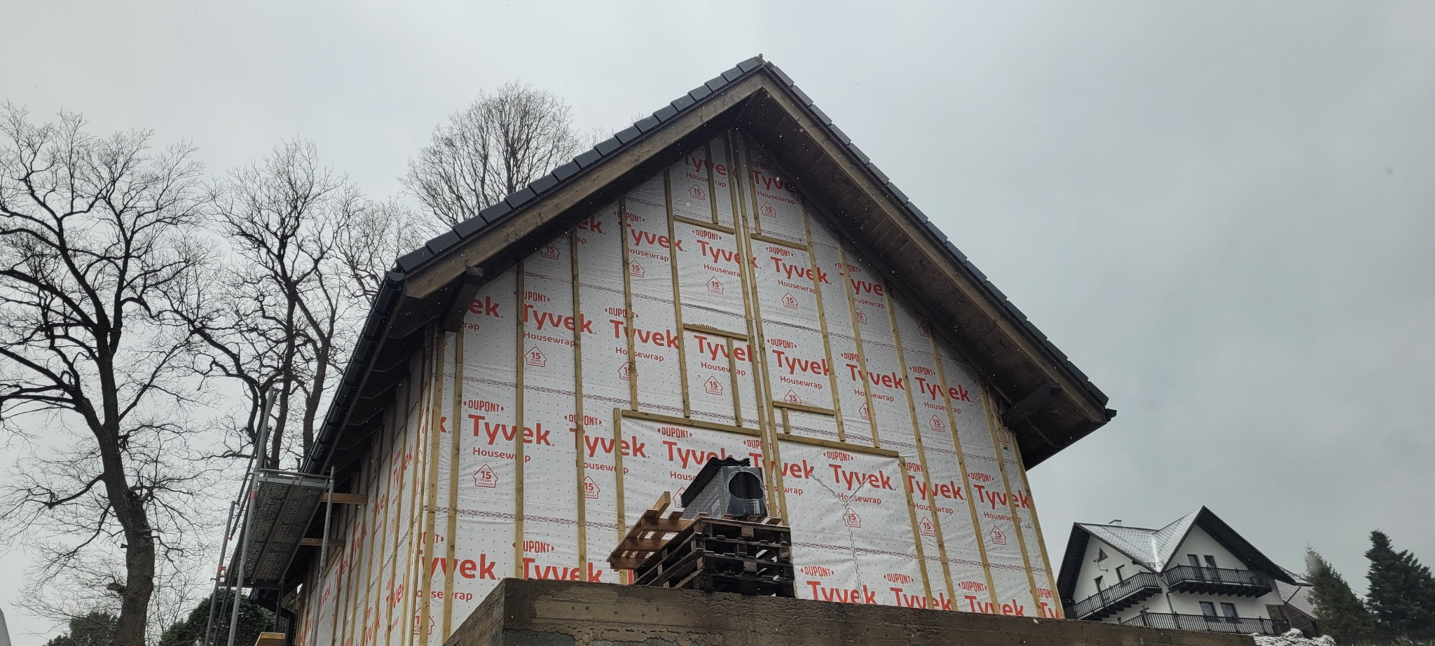 Konstrukcja domu w trakcie budowy, widoczne drewniane elementy szkieletu pokryte membraną Tyvek, na dachu ułożone dachówki, w tle drzewa bez liści i inny budynek.
