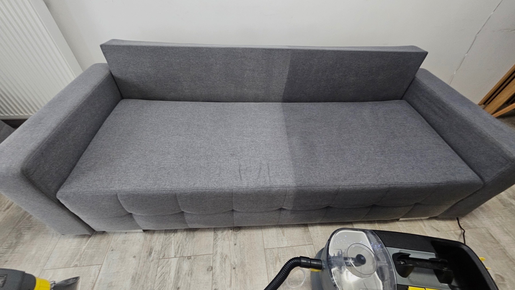 Szara kanapa z widoczną różnicą w czystości tapicerki, gdzie lewa strona jest wyraźnie ciemniejsza od wyczyszczonej prawej strony. Na podłodze widoczny odkurzacz piorący.