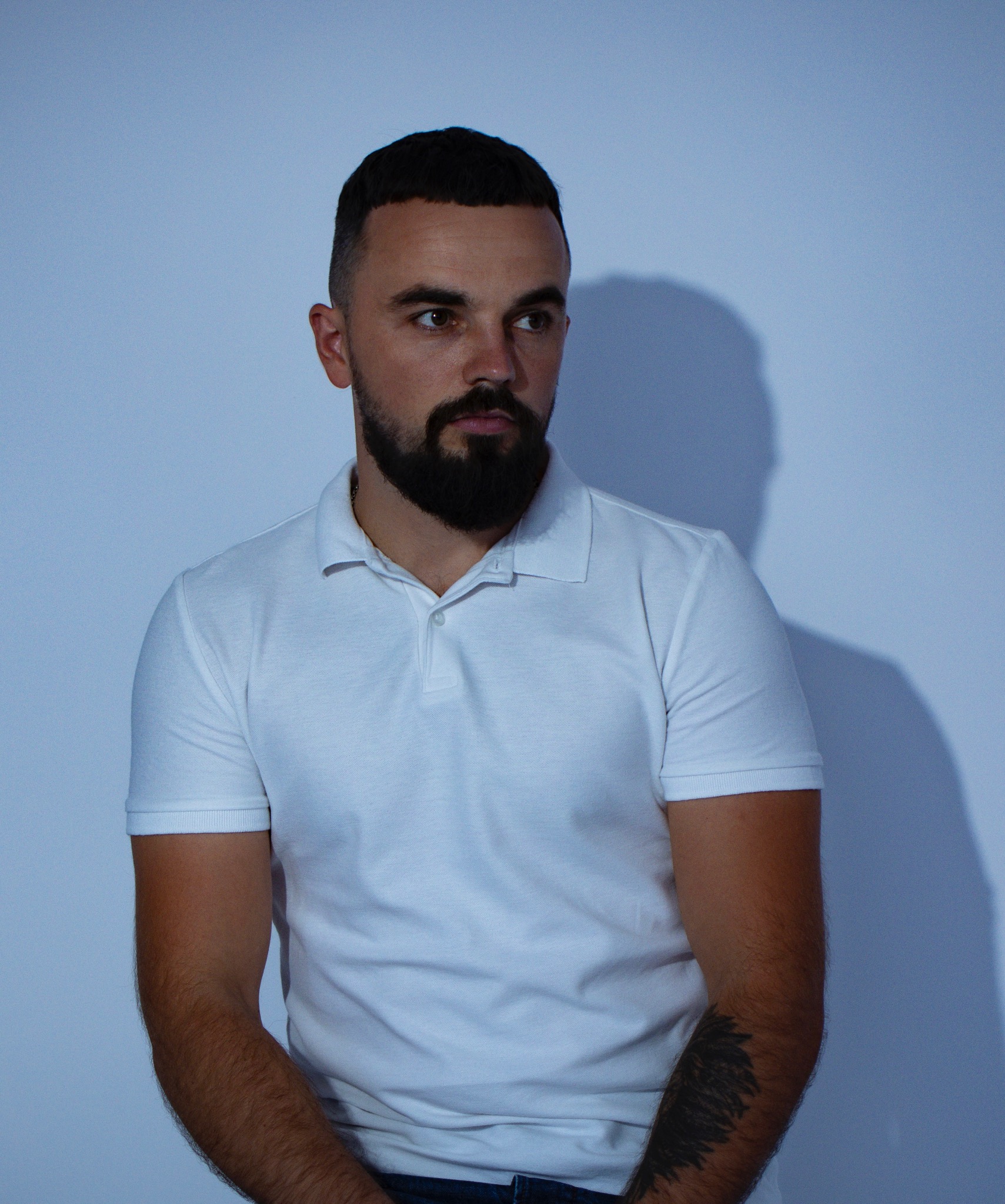 Mężczyzna z brodą w białej koszulce polo, z tatuażem na ramieniu, spogląda w bok na tle jasnoniebieskiej ściany.