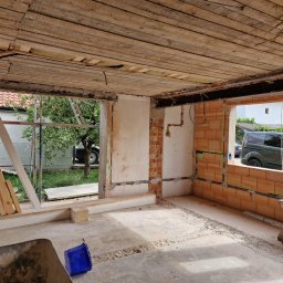 Energo-Instal Elektryk Śląsk - Wnętrze w trakcie remontu: surowy beton, odsłonięte cegły, drewniane belki stropowe, instalacja elektryczna w ścianach, widoczne okna z widokiem na zewnątrz, taczka i wiadro na podłodze.
