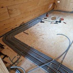 Energo-Instal Elektryk Śląsk - Szereg szarych, karbowanych rur ułożonych wzdłuż drewnianej ściany w surowym wnętrzu, zabezpieczonych metalowymi uchwytami, z widocznymi narzędziami i elementami montażowymi na podłodze.