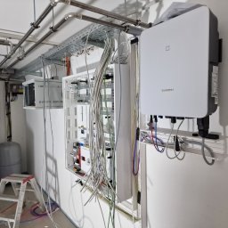 Energo-Instal Elektryk Śląsk - Wnętrze kotłowni w trakcie montażu instalacji elektrycznej: biała skrzynka rozdzielcza z przewodami, szara drabina, biały inwerter Sungrow na ścianie, rury instalacyjne pod sufitem.