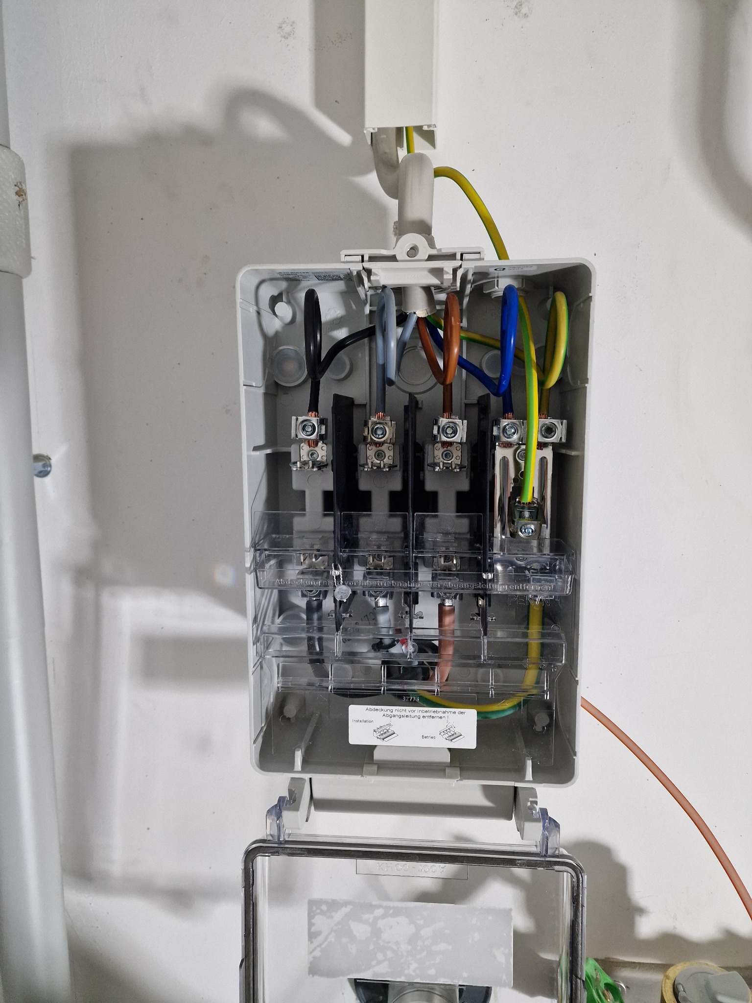 Otwarta skrzynka rozdzielcza z widocznymi przewodami o różnych kolorach (czarny, szary, brązowy, niebieski, żółto-zielony) i metalowymi zaciskami, zamontowana na białej, lekko zabrudzonej ścianie.