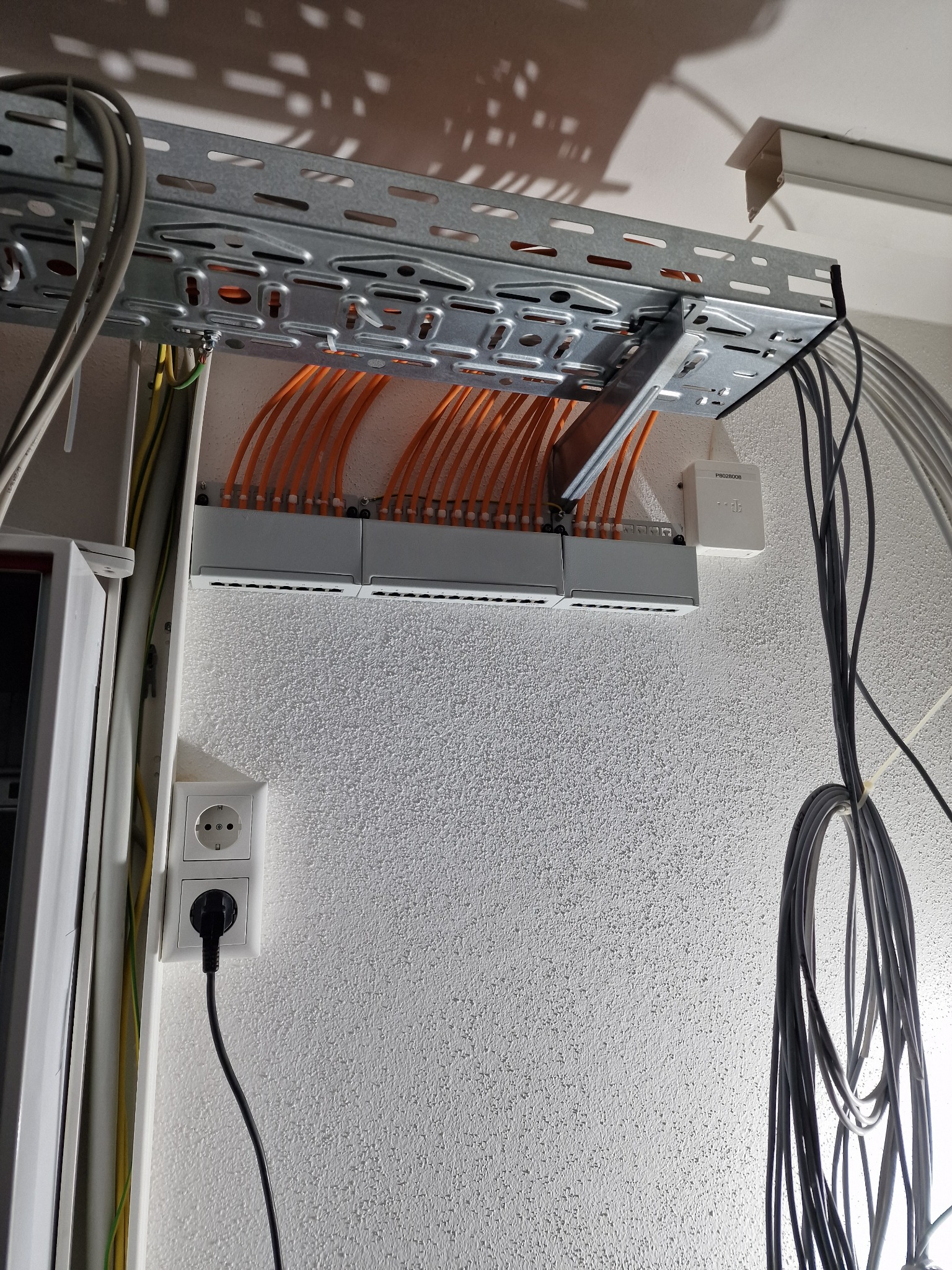 Pomarańczowe przewody sieciowe w szarej listwie montażowej, zamocowanej pod sufitem na metalowej konstrukcji. Obok widoczna puszka telekomunikacyjna i gniazdko elektryczne z podłączonym kablem.