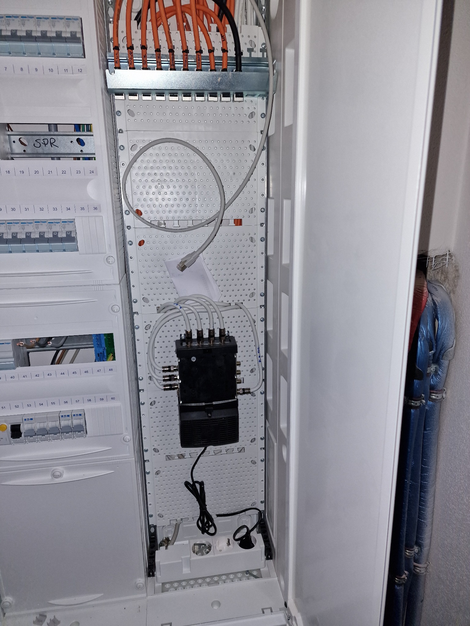 Otwarta szafa rozdzielcza z widocznymi bezpiecznikami, przewodami pomarańczowymi i szarymi kablami antenowymi, zakończonymi złączami F, podłączonymi do czarnego rozdzielacza sygnału, obok pionowy...