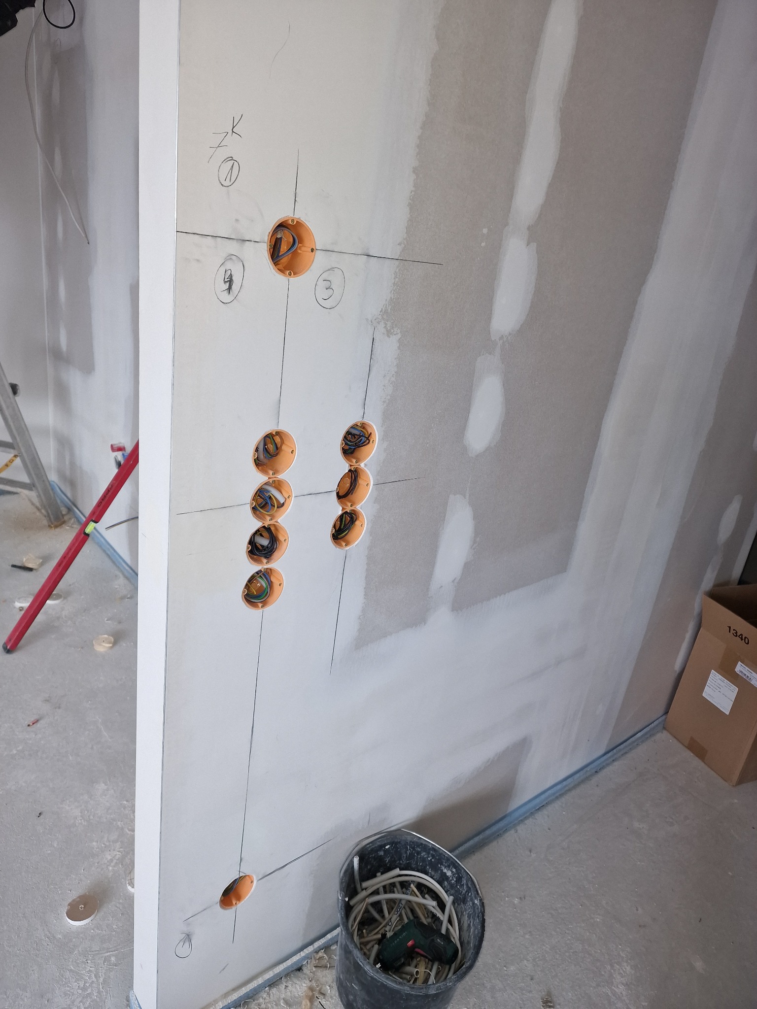 Ściana z płyt gipsowo-kartonowych z zaznaczonymi punktami i wywierconymi otworami pod puszki elektryczne, widoczne przewody wewnątrz puszek, czarne linie wyznaczające punkty, wiadro z kablami...